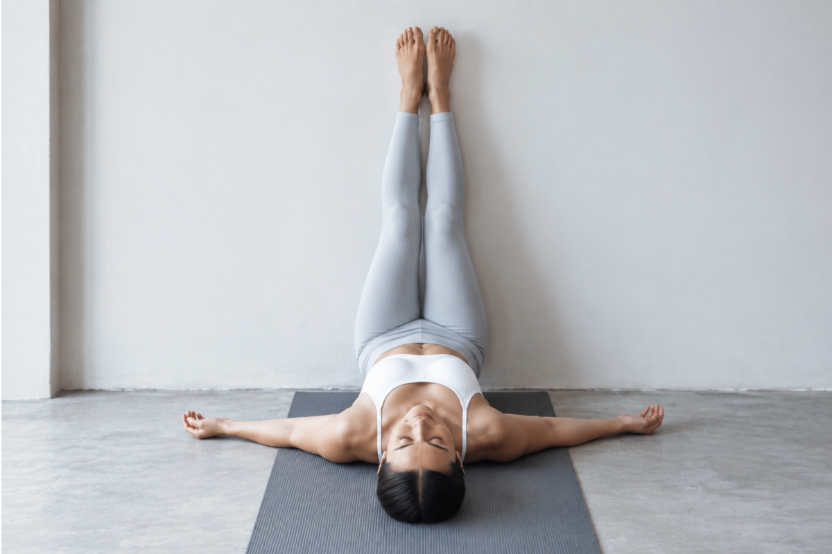 Legs up the wall. Woman lying on her back, hips close to the wall, legs extended straight up against it, arms relaxed at her sides, optional folded blanket under the hips.